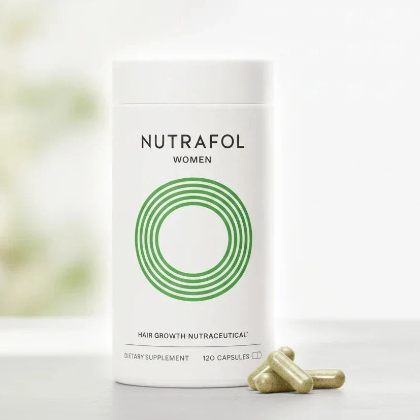 A white cylindrical bottle of Nutrafol Women dietary supplement with a green concentric circle logo, shown on a light surface with two green capsules in front. The background is a soft, blurred white and light green setting.